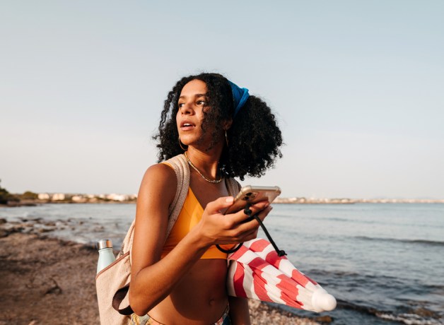Woman on beach wearing a tank top, holding her phone and looking into the distance.