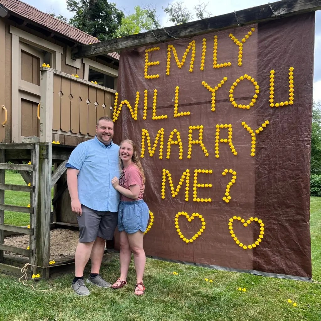 Emily and Kyle stood in front of a large piece of hanging, brown tarp with the words "Emily will you marry me?" and three hearts spelled out in yellow rubber ducks.