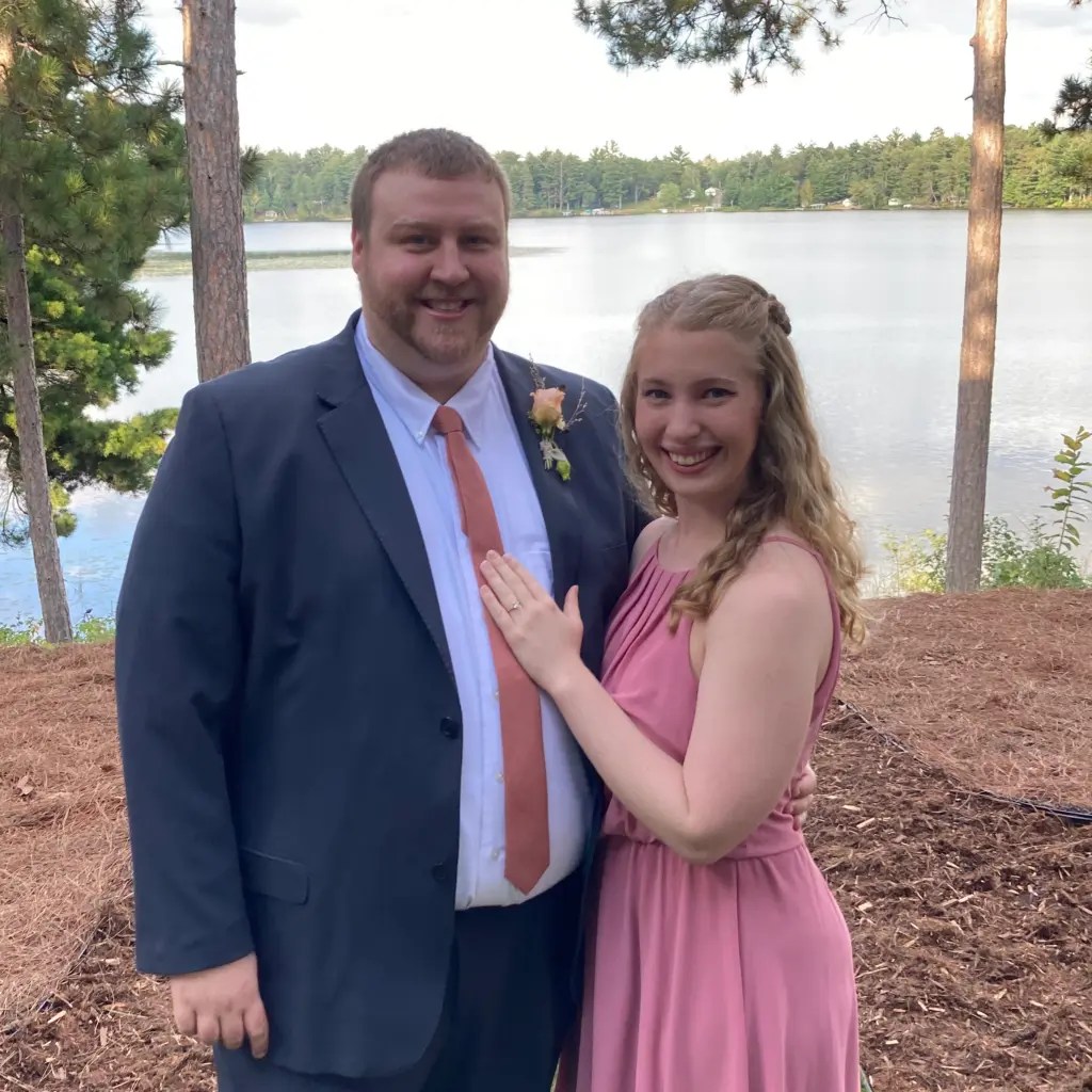 A photo of Emily and Kyle standing in front of a lake and dressed to attend a wedding with Kyle in a navy suit and Emily in a pink dress.