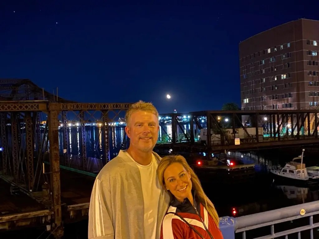 Jim and Meredith standing in front of a harbor at nighttime and smiling.