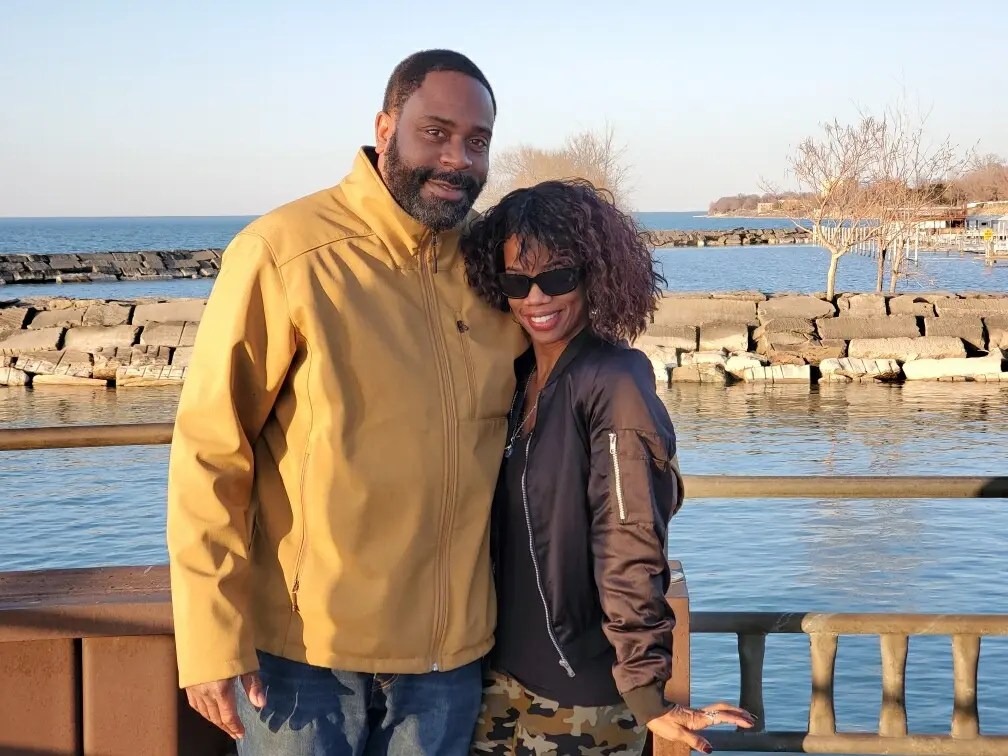 Nyla and Jeff are stood with their arms around each other and smiling in front of a harbour by the sea.