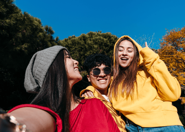 Three friends sitting together. One is wearing a yellow hoodie the other is wearing a red shirt. Another one is wearing sunglasses. It's a sunny day.