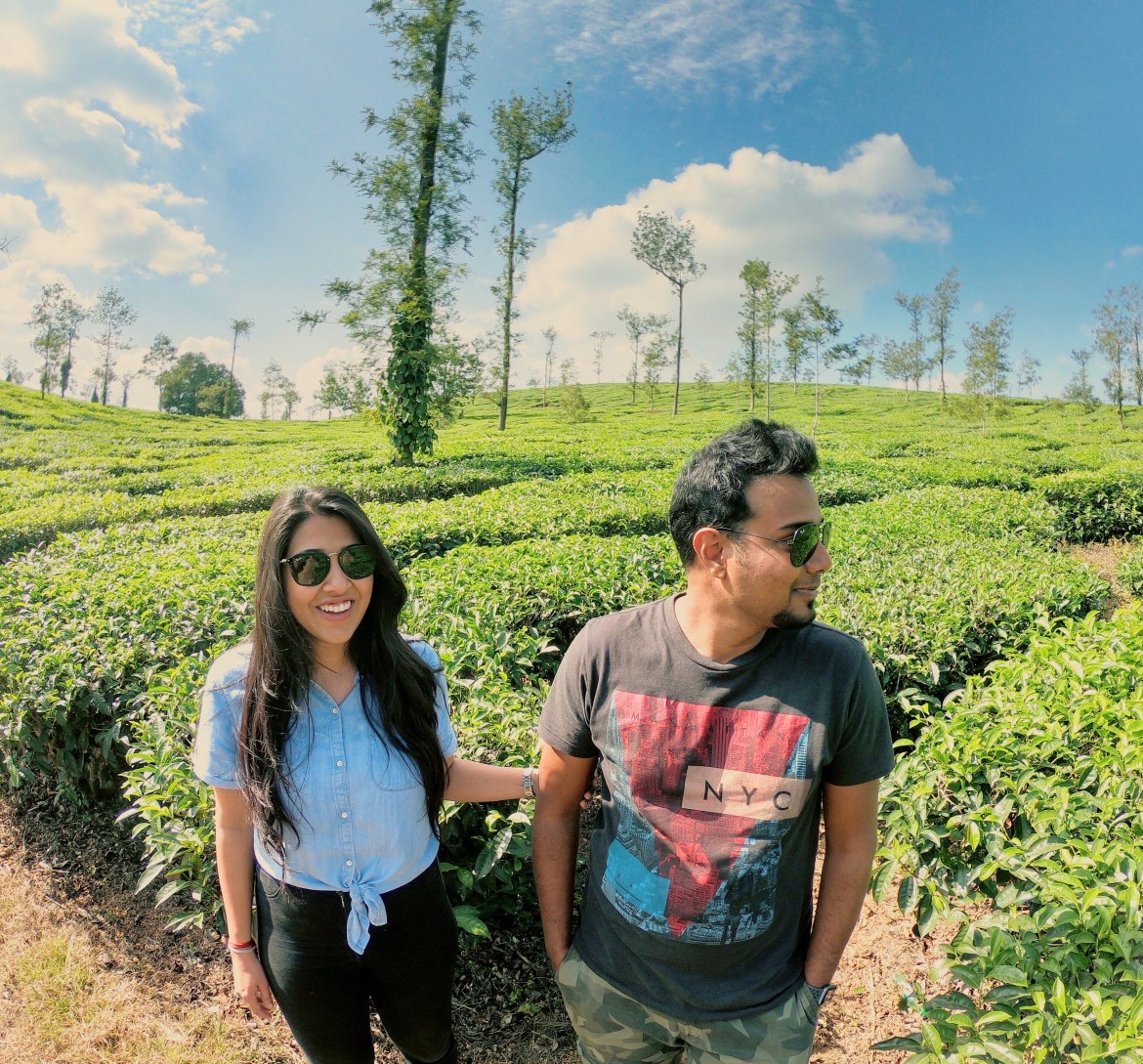 Deepti and Kishan standing together in a tea leaf field on a sunny day. Deepti is holding Kishan's arm while he looks away from the camera. 