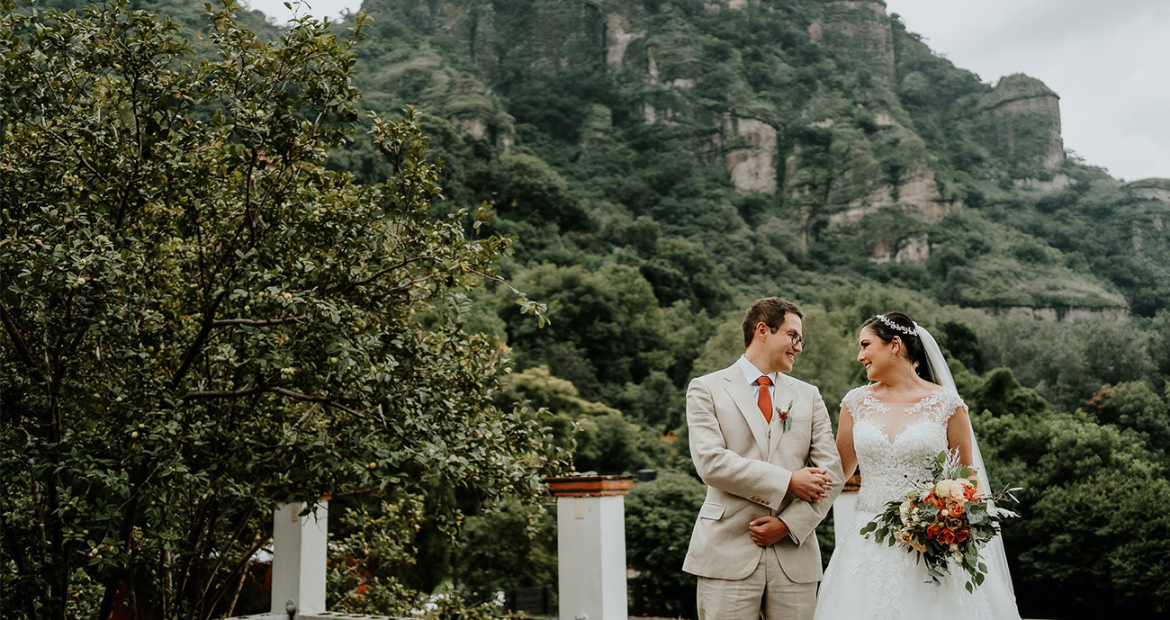Paulina and Windsor smiling at each other on their wedding day, standing in front of a beautiful, foliage-covered mountain. Windsor is wearing a cream suit and Paulina is wearing a white wedding dress and holding a bouquet of flowers.