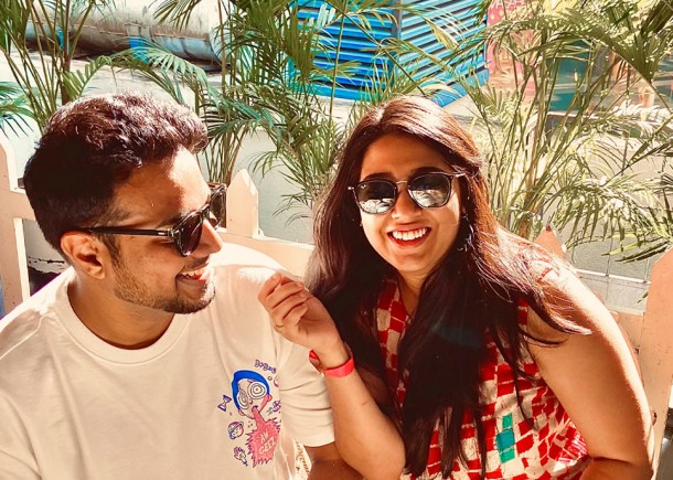 Deepti and Kishan smiling at the camera while out at a restaurant together. Kishan is wearing a white T-shirt and Deepti is wearing a bright patterned blouse. They are sat in front of greenery.