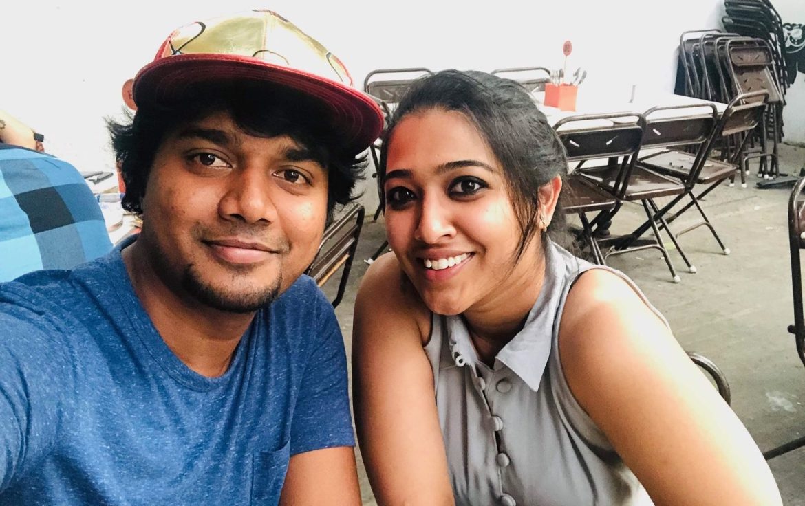 Deepti and Kishan smiling at the camera while taking a selfie together. Kishan is wearing a blue top and a hat. Deepti is wearing a grey blouse.
