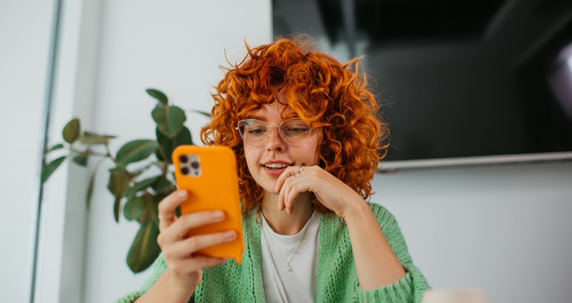 A young woman wearing a green cardigan looking at her phone which is in an orange case.