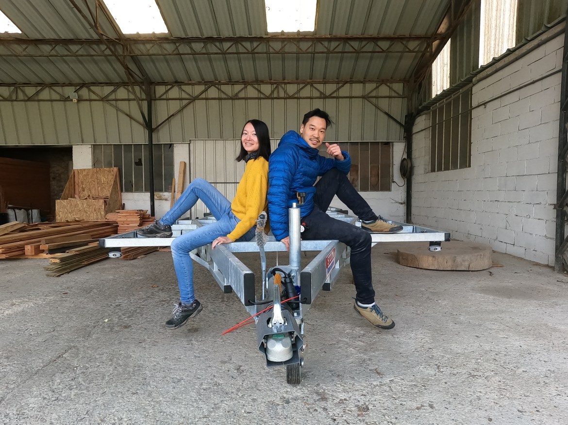 Carène and You-Liang sitting either side of their trailer. Carène is wearing a yellow jumper and You-Liang is wearing a blue jacket. 