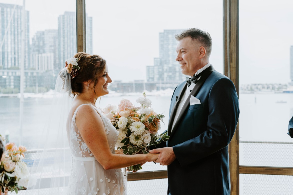 Jude and Ben are standing together at their wedding. Jude is wearing a sleeveless wedding dress and Ben is holding her hands. They're both standing in front of a big window that looks out over a city.