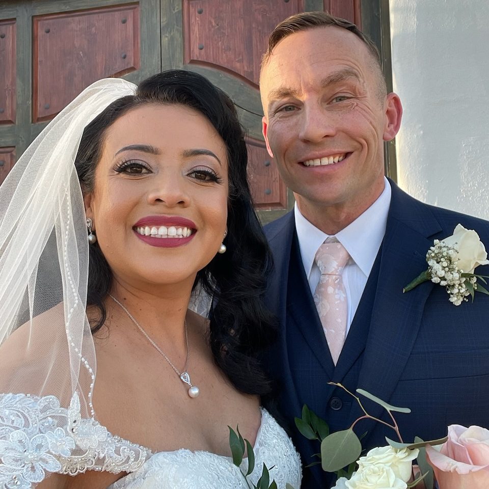 Matt and Iris are standing together on their wedding day. Both are smiling at the camera. 