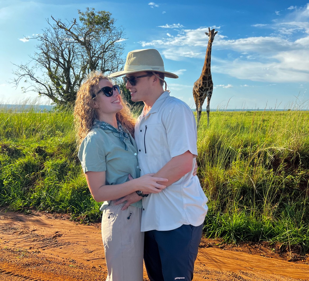 Alanna and Luke on their travels together in Africa. There is a giraffe in the background. Luke is standing with Alanna, wearing a white shirt and a hat. Alanna is wearing a buttoned blue shirt and shorts.