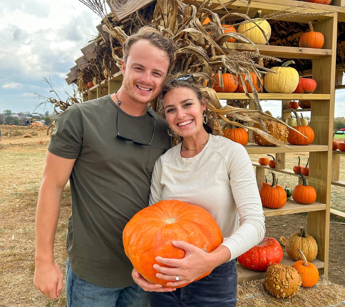 Alanna and Luke are standing together at a pumpkin patch. Alanna is holding a large pumpkin. Both are smiling at the camera.