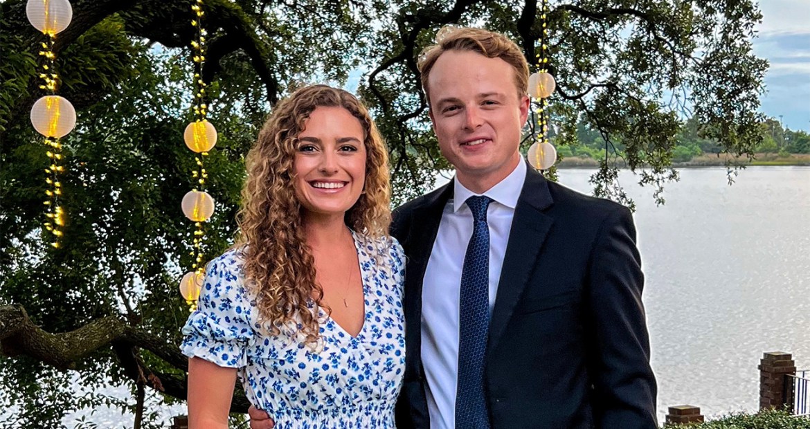Alanna and Luke are standing together in a garden dressed in formalwear. Alanna is wearing a blue and white dress. Luke is wearing a suit with a blue shirt.