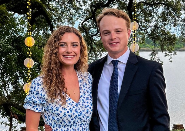 Alanna and Luke are standing together in a garden dressed in formalwear. Alanna is wearing a blue and white dress. Luke is wearing a suit with a blue shirt.