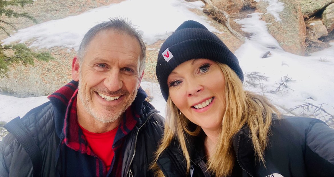 Chris and Helen are standing together in front of a snowy background. Helen is wearing a hat and both are smiling at the camera.