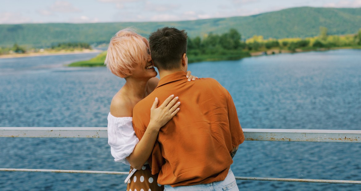 A couple is standing together looking out across a lake. The woman is holding her partner's back and whispering into their ear.