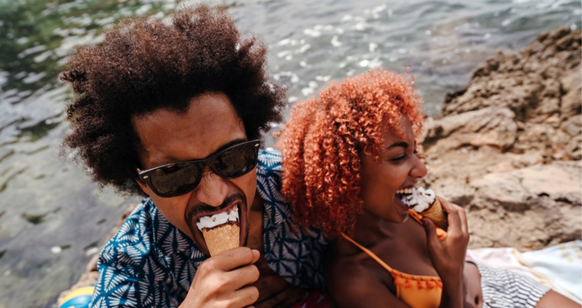 A couple sitting together on a beach eating ice creams. The man is wearing sunglasses and the woman is wearing an orange bikini top.