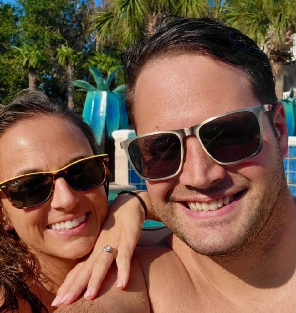 Brian and Kaci are taking a selfie together on holiday. Both are wearing sunglasses. 