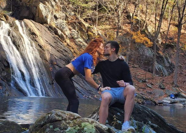 Paige and Shiloh share a kiss in front of a waterfall. Paige wears a blue top, while Shiloh is dressed in shorts.