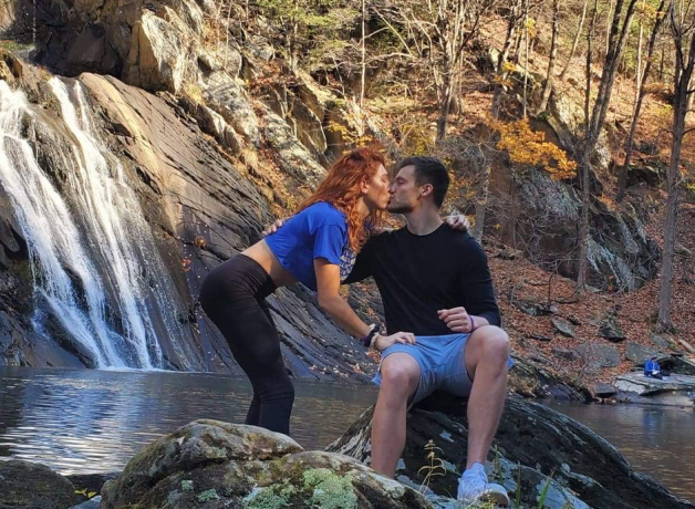 Paige and Shiloh share a kiss in front of a waterfall. Paige wears a blue top, while Shiloh is dressed in shorts.