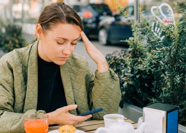 Young woman looking stressed and reflective while holding her phone at an outdoor cafe, illustrating the confusion and emotional toll that love bombing can bring in relationships.